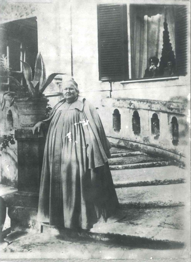 Nadine Helbig seisoo Villa Lanten pääoven portaiden alaosassa, katse kameraan ja kaiteeseen nojaten. Taustalla olevasta ikkunasta kaksi henkilöä seuraa valokuvausta.