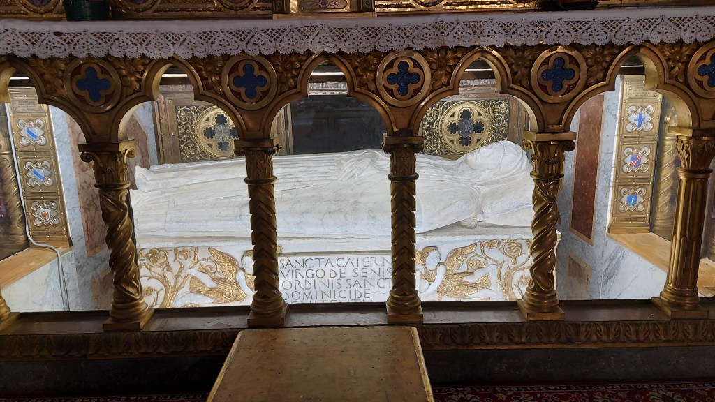 Valaistu koristeellinen hautapaikka, jonka sisällä marmoriin veistetty lepäävä ihminen kädet vatsan päällä. Veistoksen edessä kullanväriset koristeelliset pilarit.