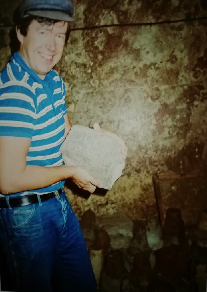 Hymyilevä mies sinivalkoisessa raitapaidassa pitää kädessään ja näyttää kameralle cippus-kiveä.